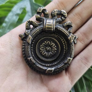 May include: An ornate bronze drawer pull with a circular design. The pull features a central floral motif, surrounded by concentric circles and decorative embellishments. The top has a bow-like design. A vintage-style hardware piece.