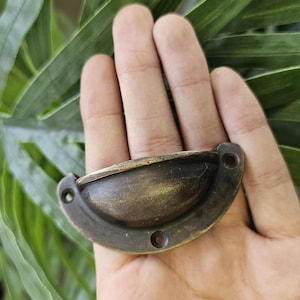 May include: Antique brass-finished drawer pull with a half-moon shape. The hardware has two mounting holes and a curved handle. The finish has a distressed look, with variations in colour from dark brown to gold.
