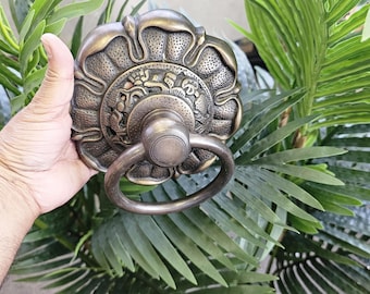 May include: Ornate bronze door knocker with a floral design and a circular handle. The knocker is held against a backdrop of green foliage, showcasing its intricate details and antique finish.