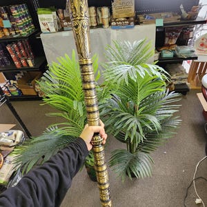 May include: An ornate, gold-colored walking stick with intricate carvings and a detailed design. The stick is held vertically, showcasing its length and decorative elements. The overall aesthetic suggests a vintage or antique style.