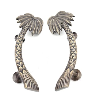 May include: Two bronze-colored palm tree door handles. Each handle is curved, with a palm frond at the top and a textured trunk. The handles are mounted on round, bronze-colored backplates, and are set against a white background.