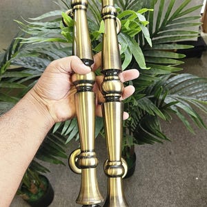 May include: Two antique brass door handles with a decorative design. The handles have a long, slender shape with ornate detailing and a warm, golden hue. The handles are held in front of a green leafy background.