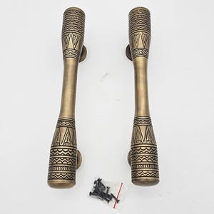 May include: Two bronze-colored door handles with intricate geometric patterns. The handles have a cylindrical shape with decorative detailing at the top and bottom. Small screws are included. The handles are designed for doors.
