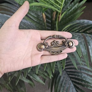 May include: An antique bronze drawer pull with decorative detailing. The pull has a curved handle with ornate flourishes and two round knobs. The hardware is held in a hand against a backdrop of green foliage.