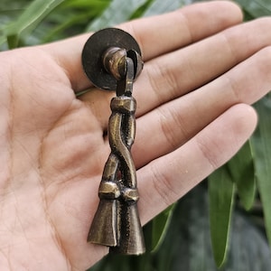 May include: A bronze cabinet pull with a decorative twisted design. The pull has a circular backplate and two tassel-like ends. The hardware is held in a hand against a backdrop of green foliage.