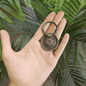 May include: A bronze cabinet pull with a circular design and a floral centre. The pull has a ring handle and is set against a backdrop of green foliage. The hardware is likely for furniture.