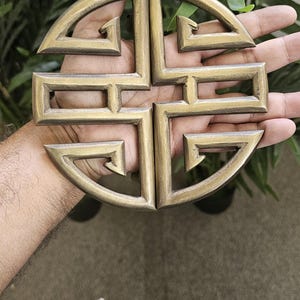 May include: A bronze-colored, four-part decorative emblem with a geometric design, held in a hand. The emblem has a circular shape with stylized, angular elements. The background includes green foliage.