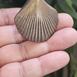May include: A small, antique brass seashell. The shell has a ridged, fan-like design and a slightly weathered appearance. The seashell is held in a hand, with green foliage in the background.
