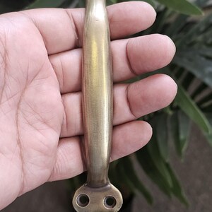 May include: A brass cabinet pull handle. The handle is held in a hand and has a rectangular shape with rounded ends. Each end has two circular holes for mounting. The handle is designed for easy gripping and pulling.