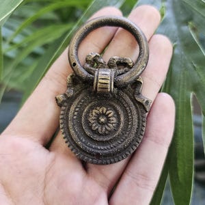 May include: An antique brass drawer pull with a circular handle and ornate, detailed design. The pull features a central floral motif and a decorative, swirling pattern. The hardware is held against a backdrop of green foliage.