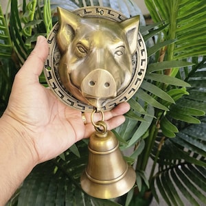 May include: A brass pig-shaped door knocker with a bell hanging below. The knocker is held in front of green foliage. The pig's face is detailed with eyes, a snout, and ears. The bell is approximately 3 inches tall.