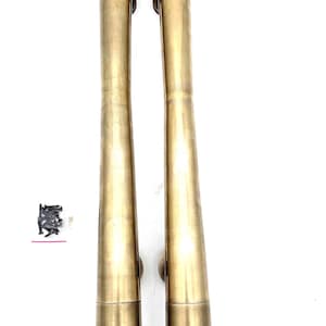 May include: Two brass-colored door pulls with a cylindrical design. Each pull has a long, slender body that tapers slightly towards the center. Small mounting hardware is included. The pulls are set against a white background.