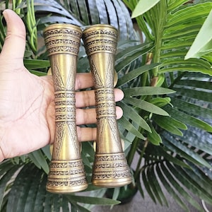 May include: Two antique brass-colored hourglass-shaped objects with intricate engraved patterns. The objects are held in a hand against a backdrop of green foliage. The design features detailed bands and geometric shapes.