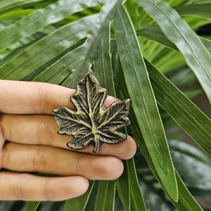 May include: A bronze-coloured maple leaf-shaped decorative object. The leaf has detailed veining and a textured surface. The object is held in a hand against a backdrop of green leaves.