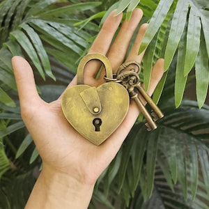 May include: A heart-shaped antique brass padlock with a keyhole and a set of keys. The lock is held in a hand, with green foliage in the background. The lock and keys have a vintage, aged appearance.