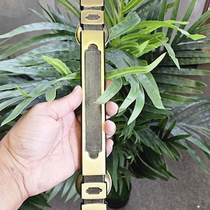 Puede incluir: Un tirador de puerta en tono latón con un diseño rectangular. El tirador presenta paneles decorativos en la parte superior e inferior, con una sección central alargada. El herraje se sostiene sobre un fondo de follaje verde.