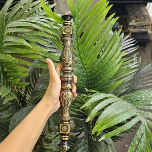 May include: Ornate, antique-style decorative column in gold and black. The column features detailed floral and geometric patterns. The object is held in front of a backdrop of green foliage, suggesting an interior design or architectural element.
