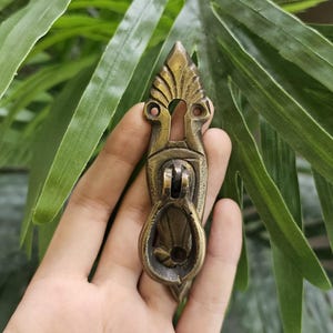May include: An antique brass-coloured cabinet pull with an ornate design. The pull features a keyhole and a decorative handle. The background is blurred, with green leaves providing a natural backdrop. This hardware piece is likely for furniture restoration.