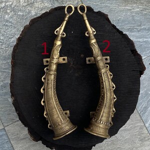 May include: Two ornate brass door handles in the shape of horns, mounted on a dark wood surface. The handles are detailed with intricate carvings and have a gold finish.