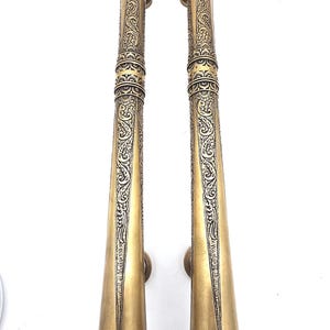 May include: Two antique brass door handles with intricate floral and geometric patterns. The handles are long, tapering, and feature small circular grips. The brass has a warm, golden hue, and the design suggests a vintage or ornate style.