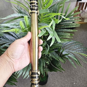 May include: A decorative bronze-colored door handle with ornate floral and geometric detailing. The handle is cylindrical with a central grip and decorative ends. The background includes green leafy plants and a gray surface.