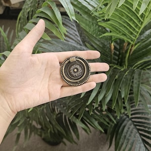 May include: A bronze-coloured drawer pull with a floral design. The pull is round with a central flower and a beaded outer edge. The background features green palm leaves.