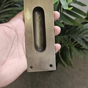 May include: A rectangular, antique brass-coloured recessed pull with a central oval finger pull. The pull has four screw holes, one in each corner. The hardware is held in a hand against a background of green foliage.