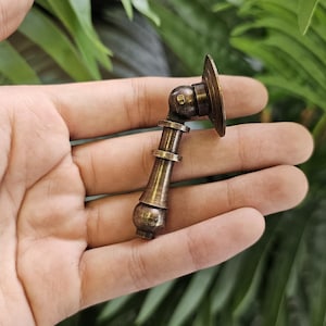 May include: A bronze-toned cabinet pull with a decorative design. The pull features a round backplate, a pivoting joint, and a cylindrical handle with decorative rings. The hardware is held in a hand against a backdrop of green foliage.