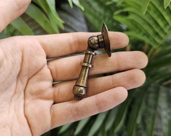 May include: A bronze-toned cabinet pull with a decorative design. The pull features a round backplate, a pivoting joint, and a cylindrical handle with decorative rings. The hardware is held in a hand against a backdrop of green foliage.