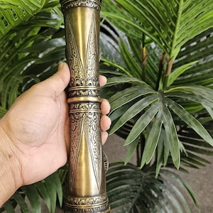 May include: A detailed, antique-style brass object with intricate patterns and designs. The cylindrical piece has a textured surface and is held against a backdrop of green foliage. The object's color is a warm, aged brass.