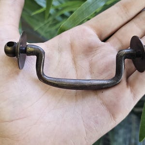 May include: Aged bronze drawer pull with a curved handle and circular backplates. The hardware is held in a hand, showing its size and design. Suitable for vintage furniture restoration or adding a classic touch to cabinets and drawers.
