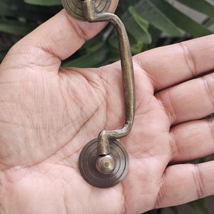 May include: Vintage brass drawer pull with a curved handle and circular backplates. The hardware has a classic aesthetic, with a warm, aged finish. The pull is designed for easy gripping and adds a touch of style to furniture.
