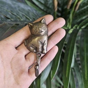 Puede incluir: Un gancho con forma de gato de color bronce. El gato está sentado con la cola enroscada. El gancho está unido a la parte inferior del cuerpo del gato. El gancho se sostiene en una mano, con hojas verdes en el fondo.