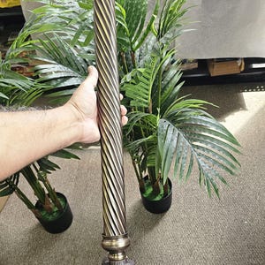 May include: Ornate bronze-colored decorative column with a spiral design. The column has decorative elements at the top and bottom. The column is held by a person's hand. The background includes green plants.