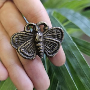 May include: A bronze-colored butterfly ring with detailed wing patterns. The ring is set against a backdrop of green leaves, highlighting the intricate design of the jewelry. The butterfly has a detailed body and antennae.