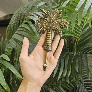 May include: A brass-colored palm tree hook, featuring a detailed palm frond design at the top and a textured trunk. The hook is held against a backdrop of lush green palm leaves, suggesting a tropical theme.