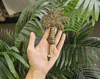 Puede incluir: Un gancho de latón con forma de palmera, con un diseño detallado de hojas de palmera y un tronco texturizado. El gancho se presenta sobre un fondo de exuberantes hojas de palmera verdes, evocando un tema tropical. Mide unos 15 cm.