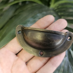 May include: A vintage-style, half-moon drawer pull with a dark bronze finish. The hardware has a curved design with two screw holes on each end and a single hole in the centre. The pull is held in a hand against a backdrop of green leaves.