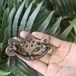 May include: An antique brass drawer pull with a decorative butterfly design. The pull features a curved handle and ornate detailing, perfect for adding a vintage touch to furniture. The hardware is held in a hand against a backdrop of green foliage.