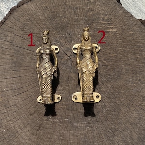 May include: Two gold-toned door handles shaped like women in flowing robes. The handles are mounted on a wooden surface. The handles are numbered 1 and 2.