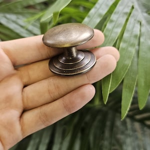 May include: A bronze cabinet knob with a rounded top and a tiered base. The hardware is held in a hand, with green foliage in the background. This decorative knob is suitable for furniture restoration or new builds.