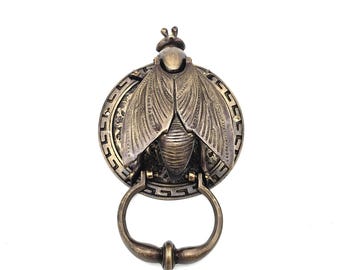 May include: Bronze-toned door knocker in the shape of a stylized insect with outstretched wings. The insect is mounted on a circular plate with a Greek key border. The knocker has a ring-shaped handle.