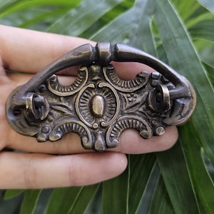 May include: Antique bronze drawer pull with ornate floral and scrollwork details. The handle is curved, and the pull has two screw holes for mounting. A decorative hardware piece for furniture restoration.