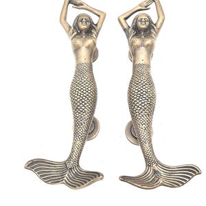 May include: Two bronze mermaid door handles. Each handle features a mermaid with arms raised, and a scaled tail. The handles are mounted on a round base. The mermaids have long hair and are facing forward.