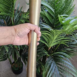 May include: A cylindrical, brass-colored metal object, possibly a torch or lamp, held by a hand. The object is long and slender, with a slightly textured surface. The background features lush green plants, suggesting an outdoor or decorative setting.