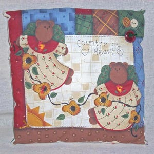 May include: Square decorative pillow with a patchwork design. Features two teddy bears wearing dresses with floral accents. The pillow has the text "Country at Heart" and is adorned with sunflower details.