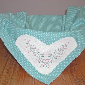 May include: A woven basket with a light blue fabric lining featuring a white heart-shaped patch. The patch is embroidered with pink flowers and green vines. The basket has a natural fiber handle.