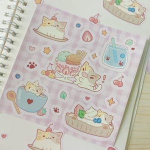 Puede incluir: Una hoja de pegatinas con gatos de dibujos animados y postres. Las pegatinas representan gatos con helados, tartas y en cuencos. El fondo es un patrón de cuadros rosas y blancos con corazones, estrellas y huellas de patas.