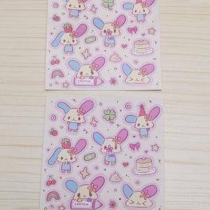 May include: Two sheets of stickers featuring a cartoon bunny with pink and blue ears, a flower, and a crayon. The stickers are on a pink background with other small cartoon images like hearts, stars, and cherries.