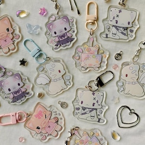 May include: A collection of clear acrylic cat-shaped keychains with various designs. The keychains feature cartoon cats in different outfits and poses, with attached metal clasps in various colors. Decorative elements like hearts and stars are scattered around.